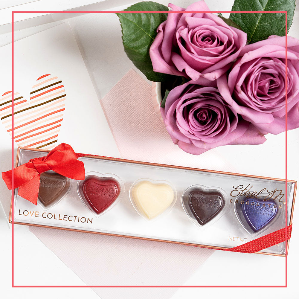 Ethel M Chocolates 5-piece Love Collection on a table with a greeting card stamped with a heart and a bouquet of pink roses.