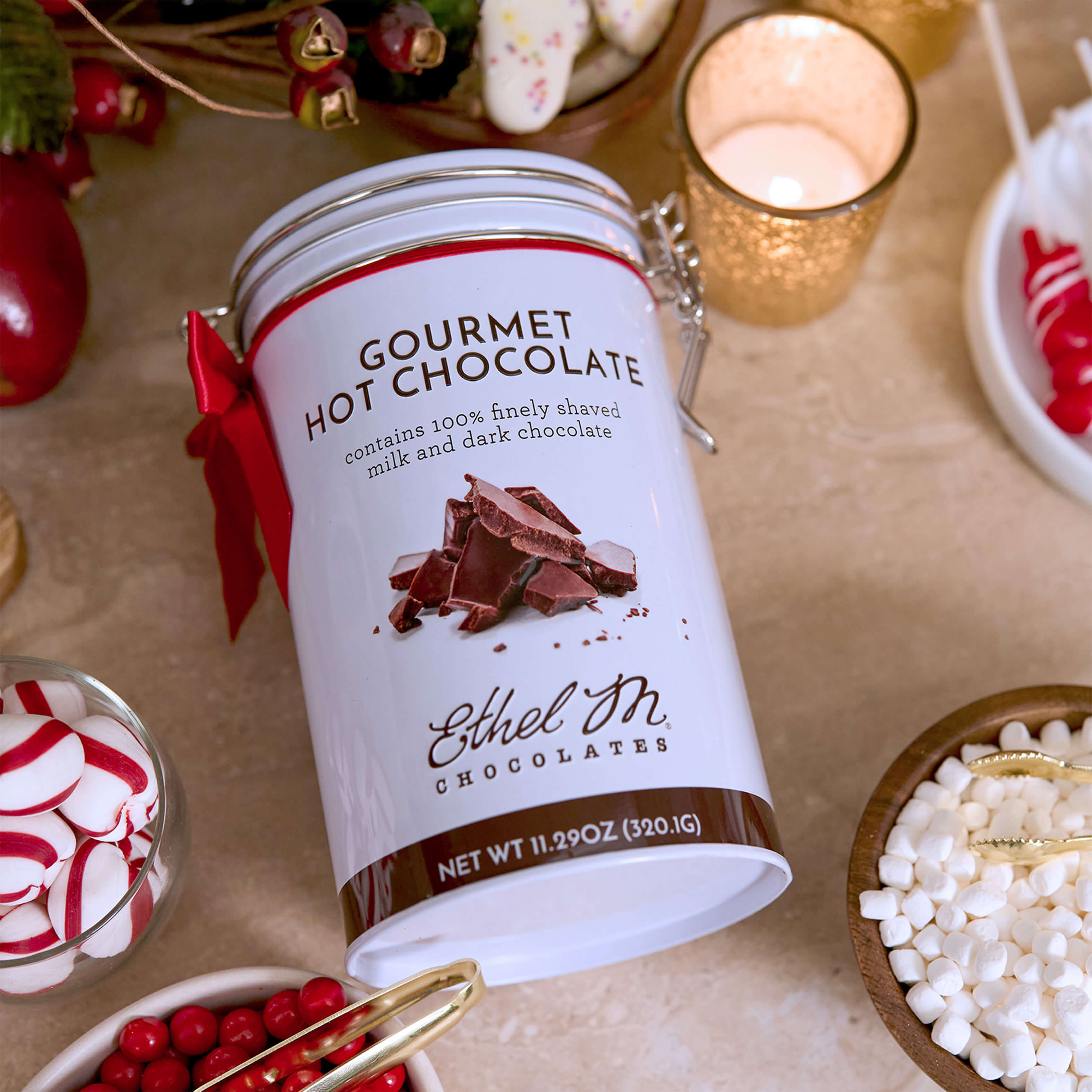 Ethel M Chocolates Gourmet Hot Chocolate Tin with Holiday Red Ribbon Lifestyle Image - Image shows the tin laying on a table surrounded by peppermints, marshmallows, and a candle