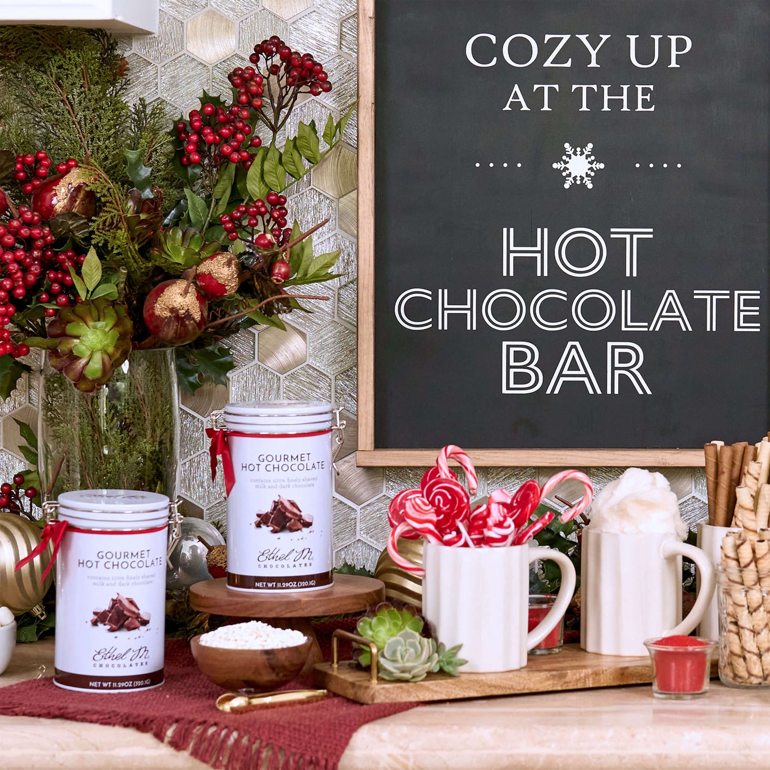 Ethel M Chocolates Gourmet Hot Chocolate Tin with Holiday Red Ribbon Lifestyle Image - Image shows two tins sitting on a bar with a mug of candy canes and a bowl of marshmallows with a sign in the background that says Cozy Up at the Hot Chocolate Bar