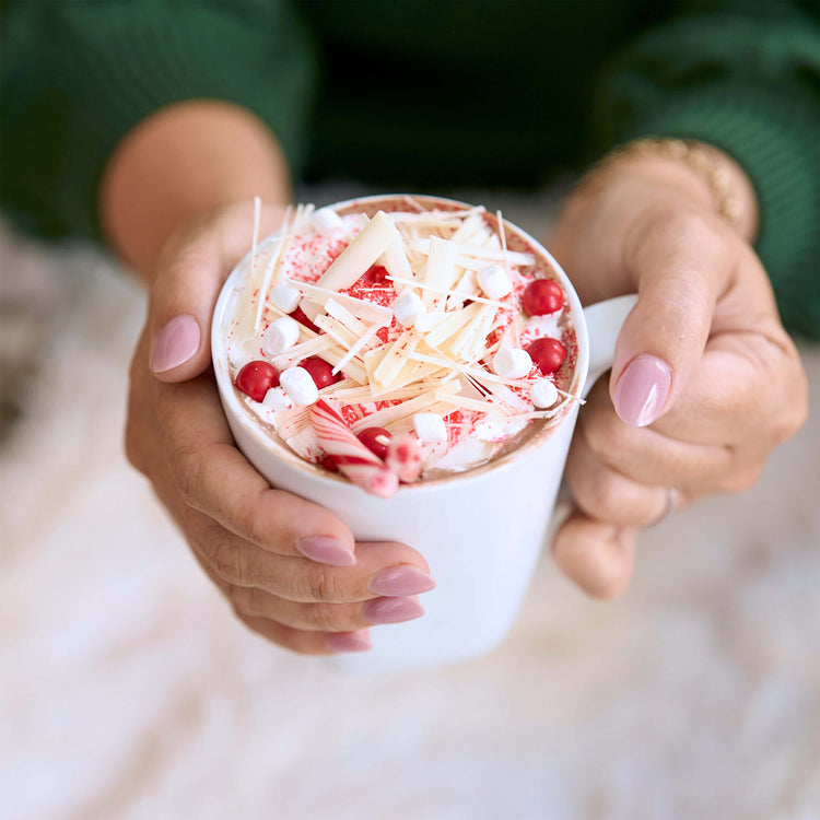 Ethel M Chocolates Gourmet Hot Chocolate Tin with Holiday Red Ribbon Lifestyle Image - Image shows a woman holding a mug of hot chocolate that is topped with marshmallows and peppermint shavings